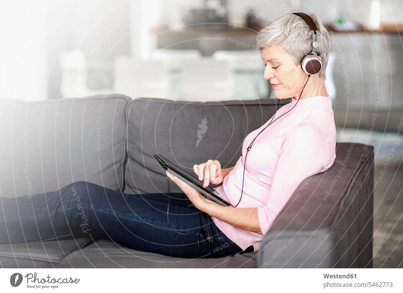 Reife Frau sitzt auf der Couch mit digitalen Tablette beim Hören von Musik mit Kopfhörern sitzen sitzend benutzen weiblich Frauen hören hoeren Tablet Computer
