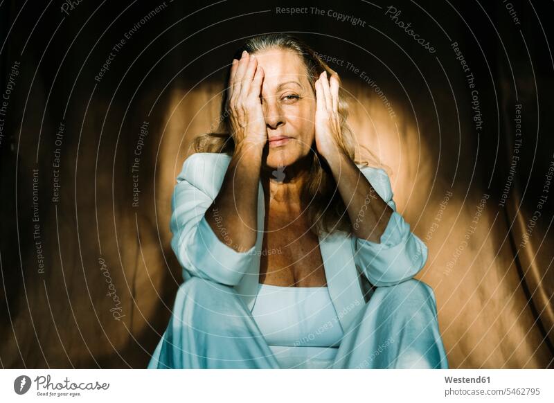 Nachdenkliche ältere Frau sitzt mit den Händen im Gesicht gegen einen braunen Vorhang Farbaufnahme Farbe Farbfoto Farbphoto Innenaufnahme Innenaufnahmen innen