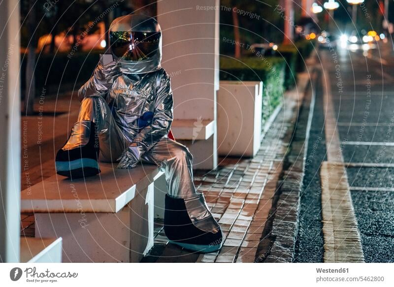 Spaceman sitzt auf Bank an einer Bushaltestelle in der Nacht hält Handy Astronaut Astronauten Haltestelle Haltestellen Bushaltestellen Straße Strassen Straßen