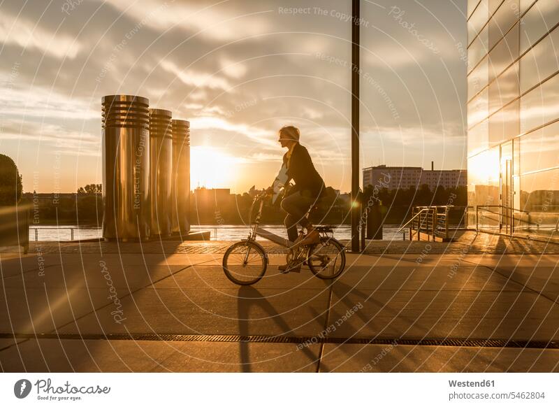 Ältere Frau fährt bei Sonnenuntergang am Flussufer mit dem Stadtrad Sonnenuntergänge Fahrrad Bikes Fahrräder Räder Rad fahren Stimmung stimmungsvoll Atmosphäre