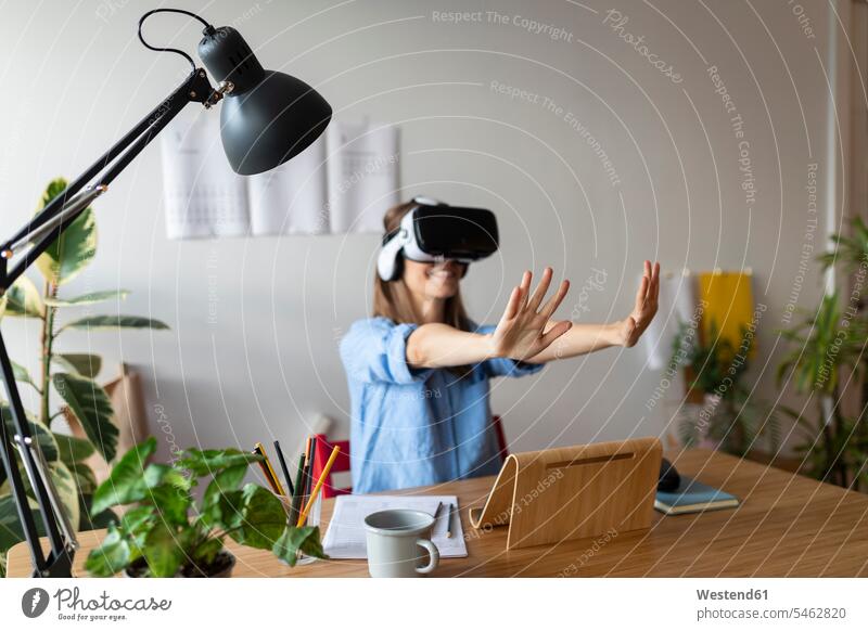 Junge Frau schaut durch einen Virtual-Reality-Simulator und gestikuliert, während sie im Home-Office sitzt Farbaufnahme Farbe Farbfoto Farbphoto Spanien