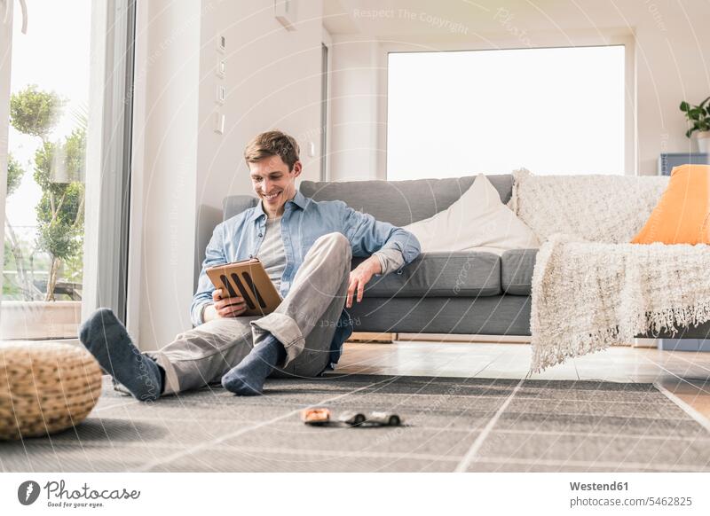Mittelgroßer erwachsener Mann sitzt auf dem Boden und benutzt ein digitales Tablett Auto Autos Spielzeugautos Teppiche entspannen relaxen Lektüre sitzend daheim