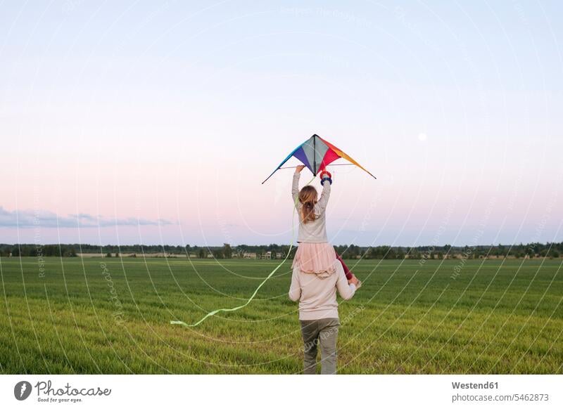 Mann trägt Tochter, die einen Drachen auf den Schultern hält, während er bei Sonnenuntergang über eine Graslandschaft geht Farbaufnahme Farbe Farbfoto Farbphoto