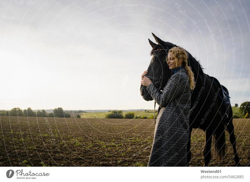 Liebevolle junge Frau mit Pferd auf einem Feld auf dem Land abends freuen geniessen Genuss Glück glücklich sein glücklichsein zufrieden stehend steht