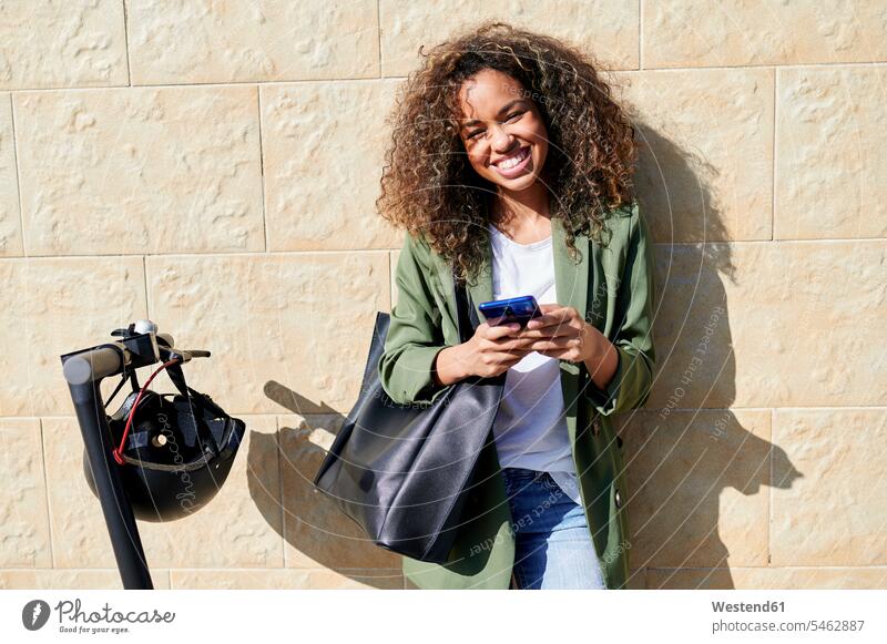 Fröhliche junge Frau hält ein Smartphone in der Hand, während sie mit einem Elektroroller an der Wand steht Farbaufnahme Farbe Farbfoto Farbphoto Außenaufnahme