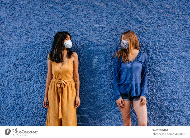 Freundinnen mit Masken, die sich gegenseitig ansehen und dabei Abstand zur blauen Mauer in der Stadt halten Farbaufnahme Farbe Farbfoto Farbphoto Spanien