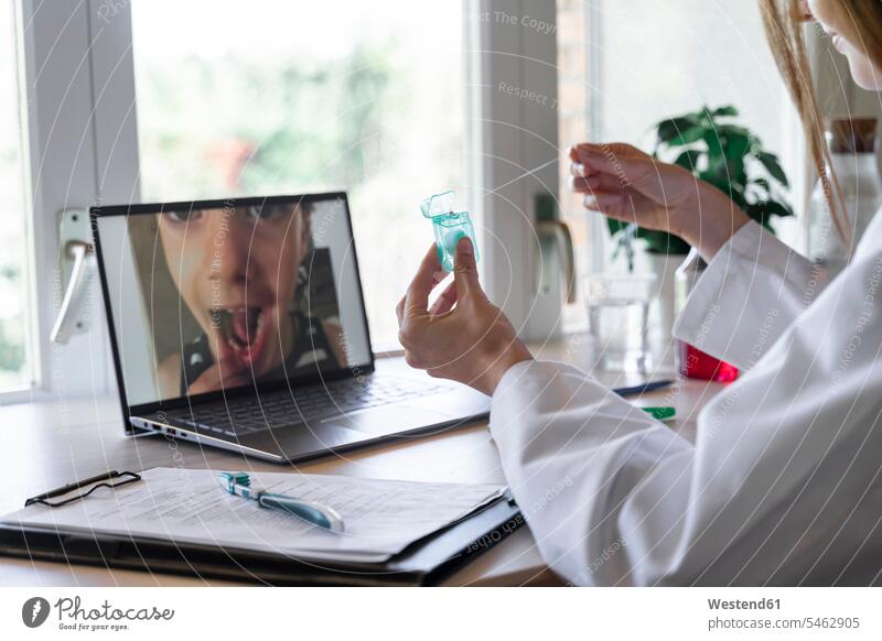 Junge Zahnärztin zeigt Patientin Zahnseide per Videoanruf auf Laptop zu Hause Farbaufnahme Farbe Farbfoto Farbphoto Innenaufnahme Innenaufnahmen innen drinnen