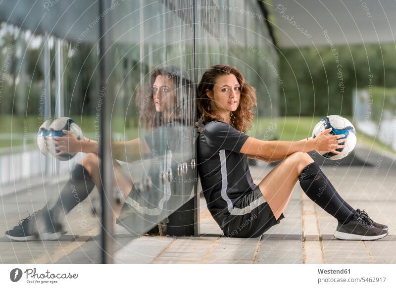 Fußballspielerin hält Ball, während sie auf einem Fußweg an einer Glaswand sitzt Farbaufnahme Farbe Farbfoto Farbphoto Deutschland Außenaufnahme außen draußen