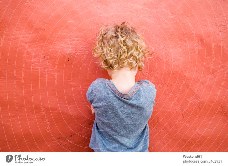 Rückenansicht eines kleinen Jungen, der vor einer roten Wand steht T-Shirts allein vereinsamt zurueckgezogen zurückgezogen gefühlvoll Emotionen Empfindung