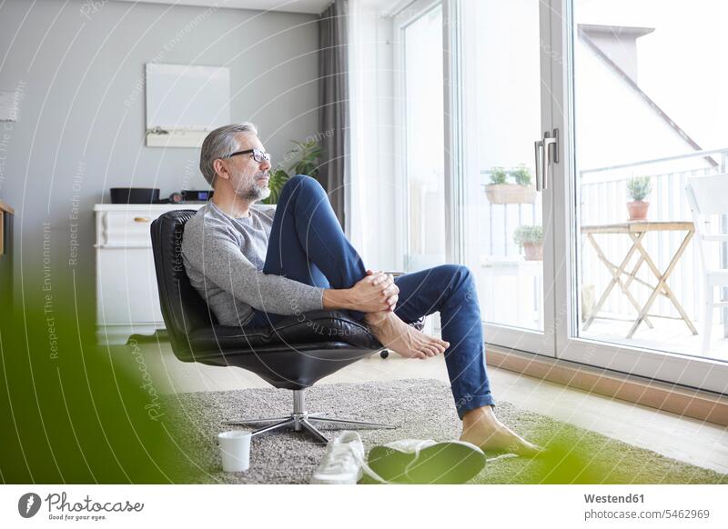 Reifer Mann entspannt sich auf einem Ledersessel in seinem Wohnzimmer und schaut aus dem Fenster Wohnraum Wohnung Wohnen Wohnräume Wohnungen Männer männlich