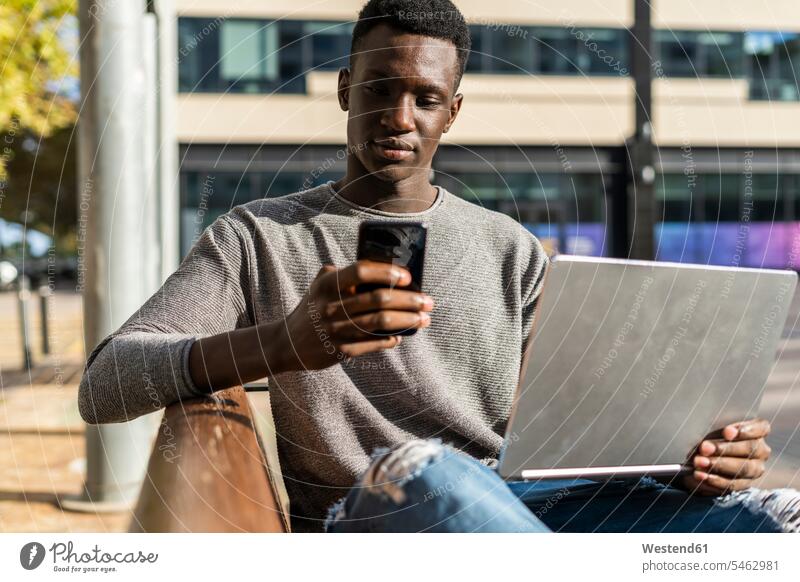 Junger Mann sitzt auf einer Bank in der Stadt, mit Laptop und Smartphone Notebook Laptops Notebooks sitzen sitzend Sitzbänke Bänke Sitzbank iPhone Smartphones