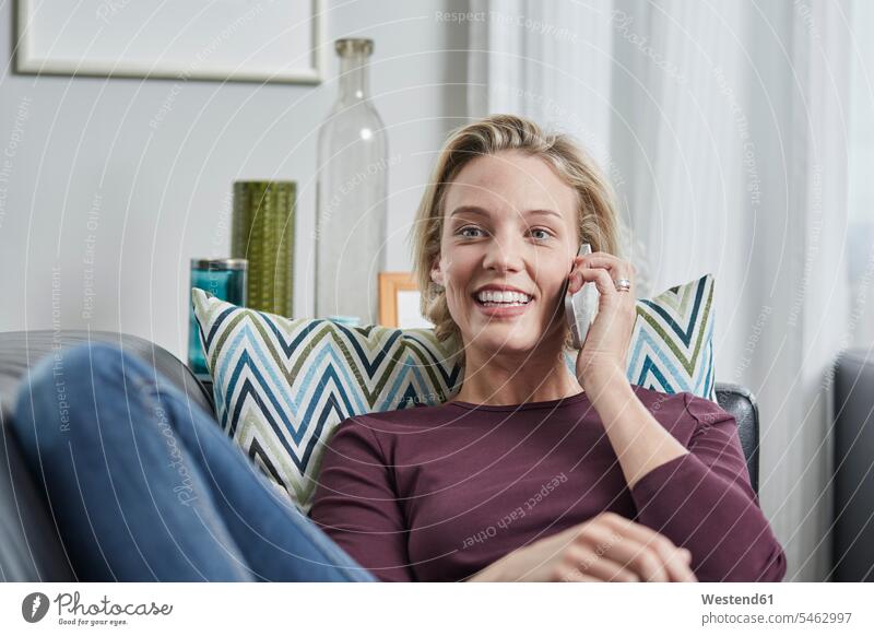 Lächelnde junge Frau am Handy liegend auf der Couch zu Hause Sofa Couches Liege Sofas Zuhause daheim lächeln Mobiltelefon Handies Handys Mobiltelefone liegt