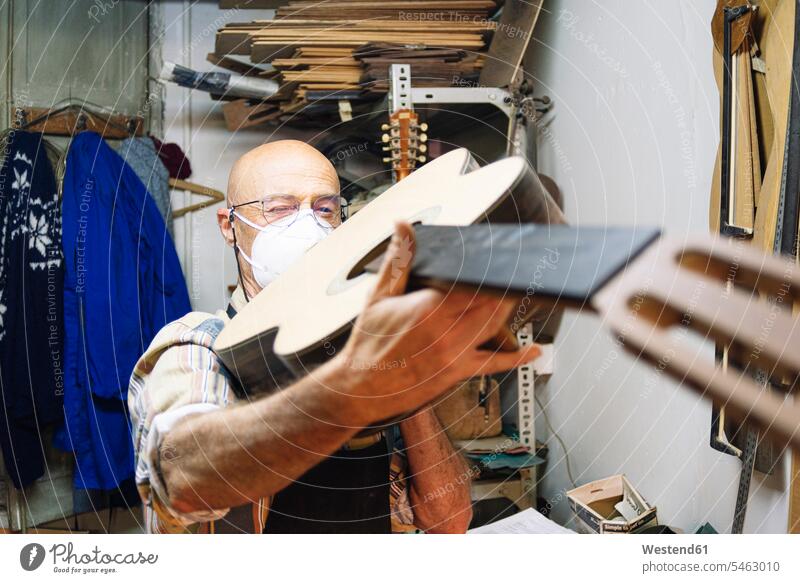 Handwerker mit Gesichtsmaske untersucht Gitarre im Stehen in der Werkstatt Farbaufnahme Farbe Farbfoto Farbphoto Innenaufnahme Innenaufnahmen innen drinnen Tag