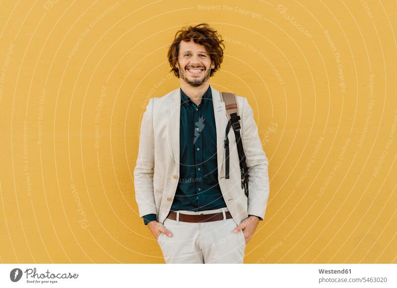 Porträt eines lachenden Geschäftsmannes mit Rucksack vor gelber Wand stehend geschäftlich Geschäftsleben Geschäftswelt Geschäftsperson Geschäftspersonen