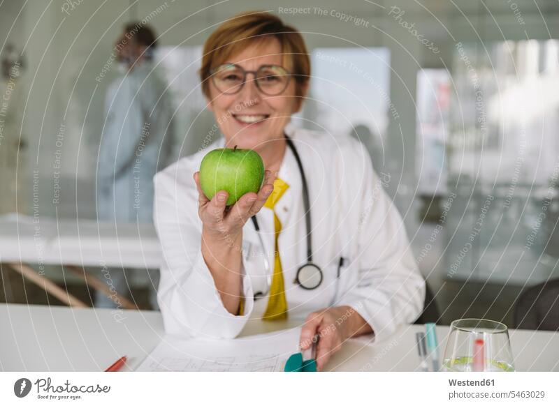 Arzt sitzt am Schreibtisch und hält einen Apfel Job Berufe Berufstätigkeit Beschäftigung Jobs Glasscheiben Gesundheit Gesundheitswesen medizinisch Stethoskope