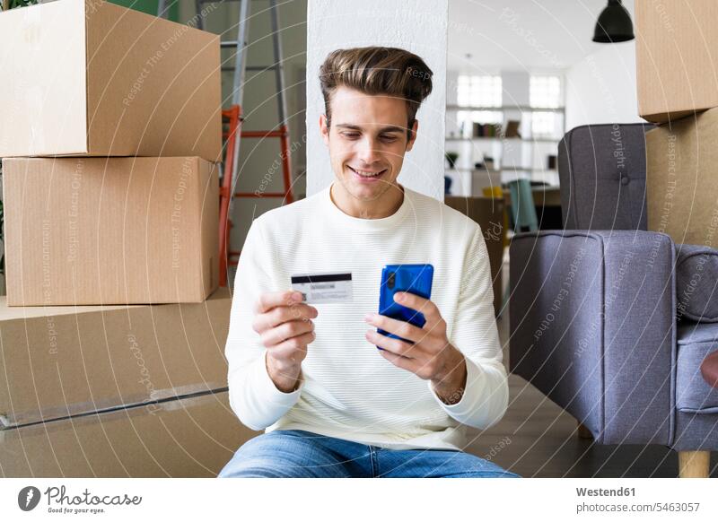 Lächelnder junger Mann, der beim Einzug in ein neues Haus per Handy mit Kreditkarte bezahlt Farbaufnahme Farbe Farbfoto Farbphoto Innenaufnahme Innenaufnahmen