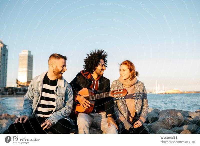 Drei glückliche Freunde mit Gitarre sitzen im Freien an der Küste Sonnenuntergang Glück glücklich sein glücklichsein sitzend sitzt Kueste Kuesten Küsten