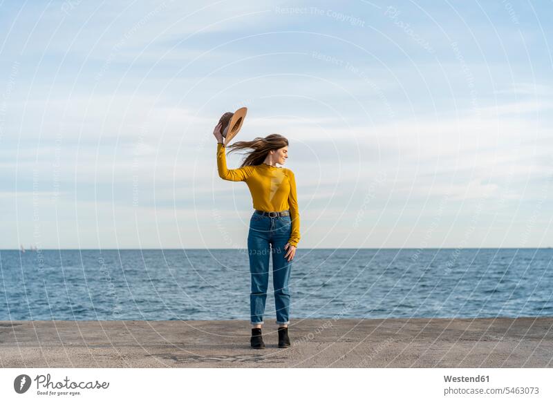 Junge Frau mit geschlossenen Augen hält Sonnenhut und genießt gegen das Meer Farbaufnahme Farbe Farbfoto Farbphoto Freizeitbeschäftigung Muße Zeit Zeit haben