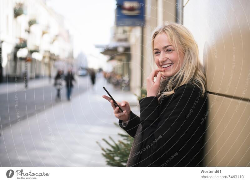Porträt einer lächelnden jungen Frau mit Handy in der Stadt blond blonde Haare blondes Haar Mantel Mäntel Jacke Drahtlose Technologie drahtlose Verbindung