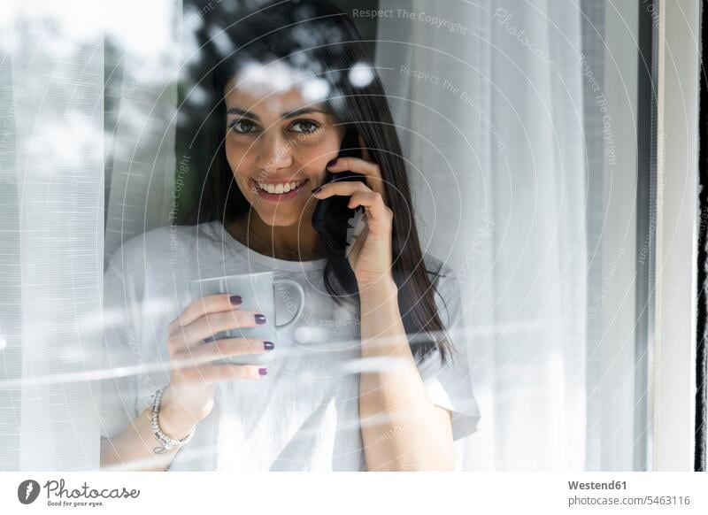 Porträt einer lächelnden jungen Frau am Handy hinter einer Fensterscheibe Leute Menschen People Person Personen Alleinstehende Alleinstehender Singles