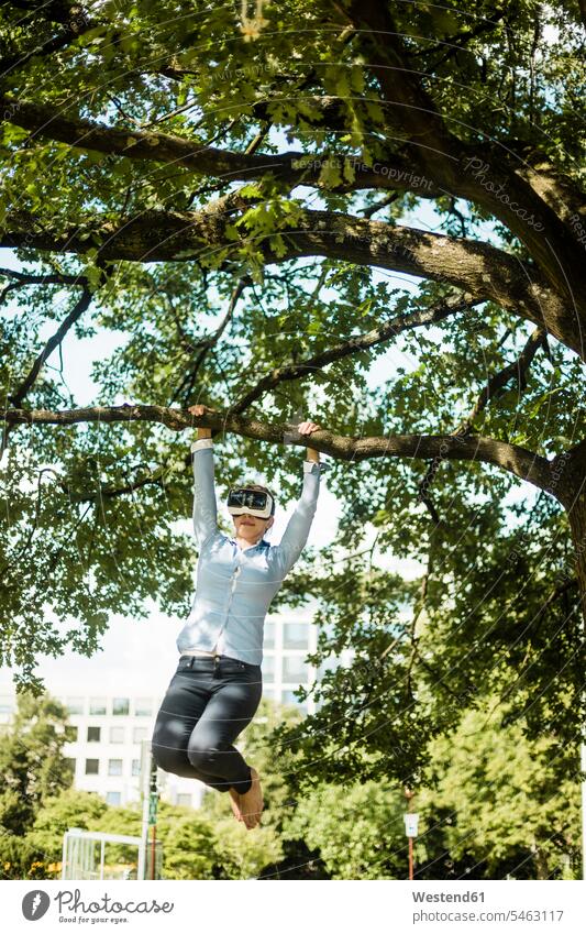 Frau im Stadtpark hängt am Ast eines Baumes und trägt eine VR-Brille Virtual Reality Brille Virtual-Reality-Brille Virtual Reality-Brille VR Brille betrachten