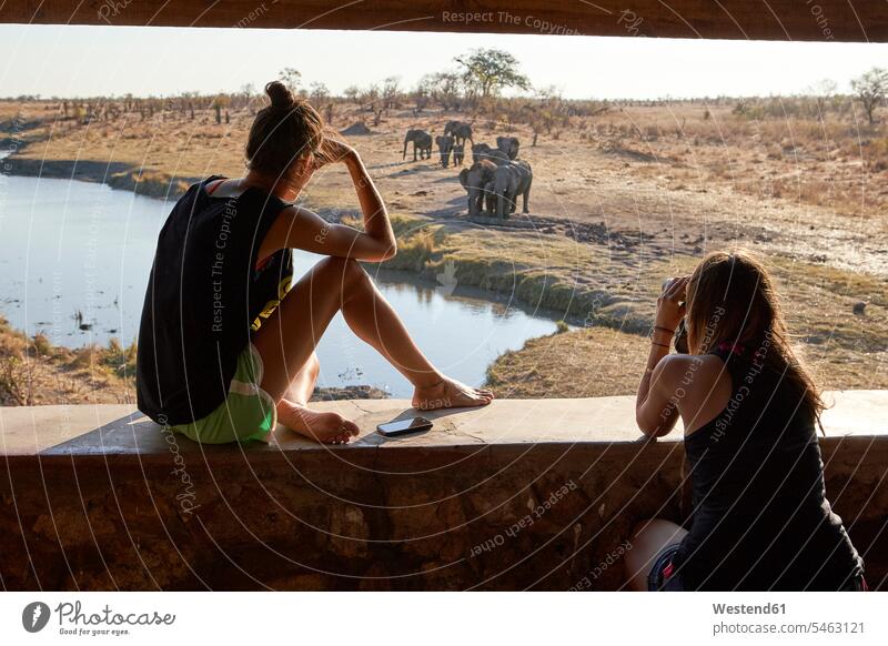 Zwei Frauen beobachten eine Elefantenherde im Fluss von einem Aussichtspunkt aus, Hwange-Nationalpark, Simbabwe Leute Menschen People Person Personen erwachsen