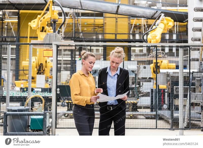 Zwei Frauen besprechen einen Plan in einer Fabrikhalle mit einem Industrieroboter weiblich Pläne Industriehallen Fabrikhallen Fabriken Roboter diskutieren