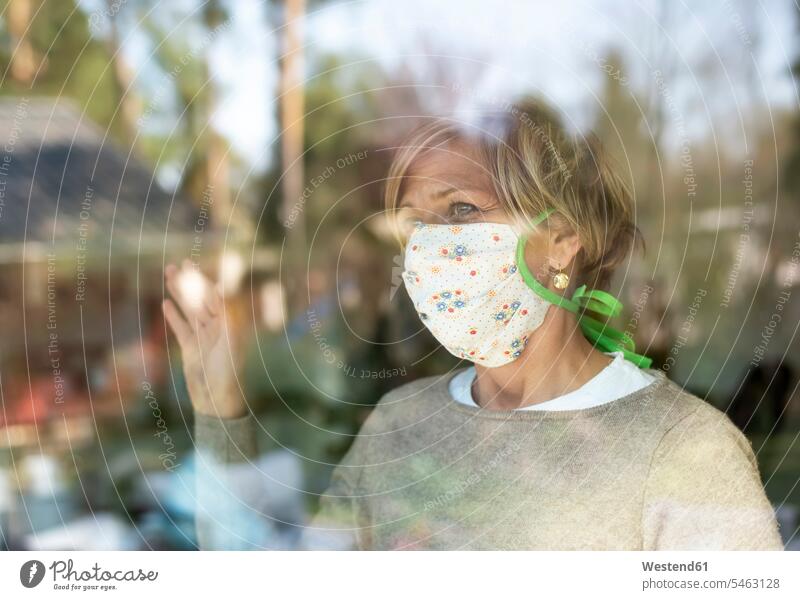 Reife Frau mit Gesichtsmaske, die während der COVID-19-Pandemie durch Fensterglas schaut Farbaufnahme Farbe Farbfoto Farbphoto Zuhause Häusliches Leben