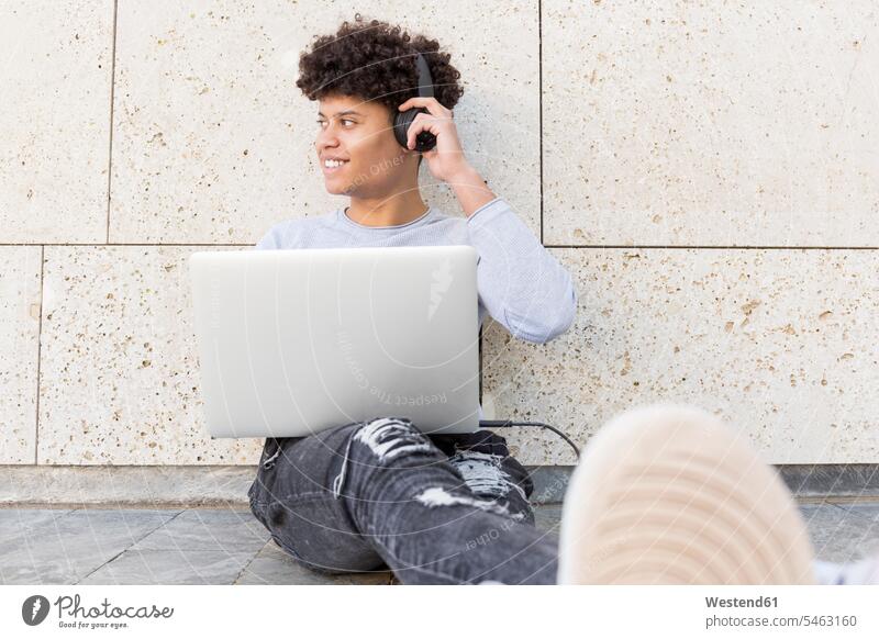 Lächelnder junger Mann mit Kopfhörern am Laptop in der Stadt Leute Menschen People Person Personen 1 Ein ein Mensch eine nur eine Person single erwachsen