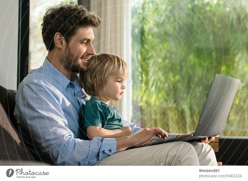 Vater und Sohn schauen sich zu Hause auf der Couch einen Laptop an ansehen sitzen sitzend sitzt Notebook Laptops Notebooks Sofa Couches Liege Sofas Söhne Papas