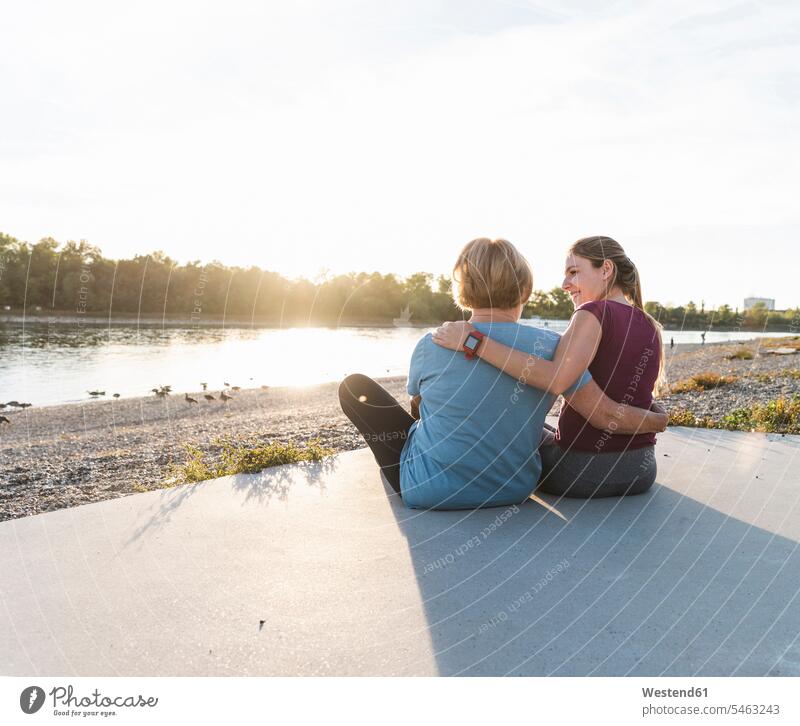 Großmutter und Enkelin sitzen nach dem Training am Fluss und beobachten den Sonnenuntergang Quality Time Erholung erholen Ganzkörperansicht Ganzansicht
