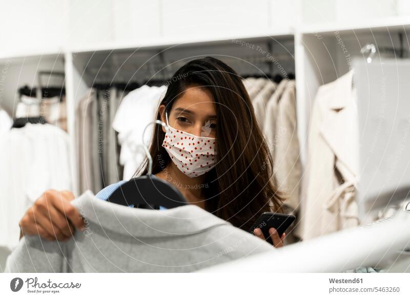 Frau mit Schutzmaske beim Betrachten von Kleidung im Einkaufszentrum Farbaufnahme Farbe Farbfoto Farbphoto Innenaufnahme Innenaufnahmen innen drinnen Tag