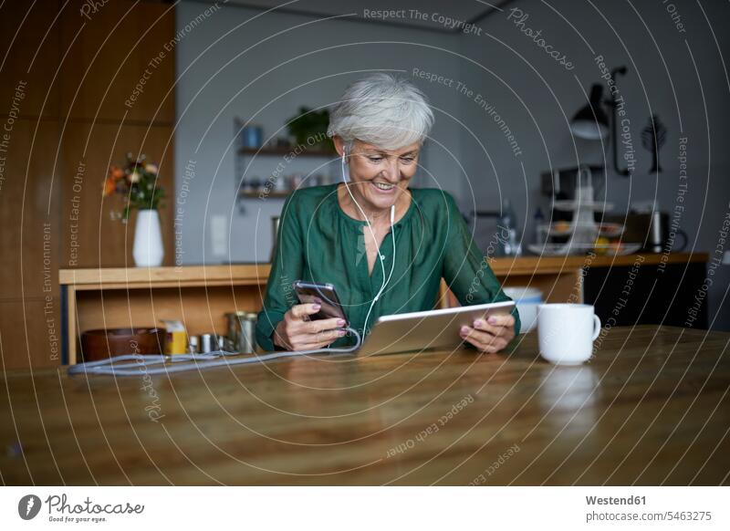Ältere Frau hört Musik, während sie zu Hause ein Smartphone und ein digitales Tablet benutzt Farbaufnahme Farbe Farbfoto Farbphoto Innenaufnahme Innenaufnahmen