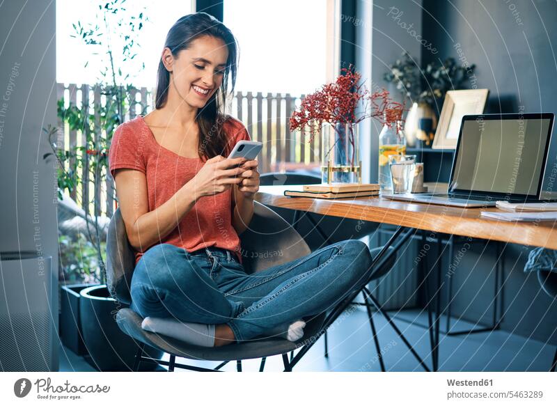 Unternehmerin benutzt Smartphone, während sie im Heimbüro auf einem Stuhl am Schreibtisch sitzt Farbaufnahme Farbe Farbfoto Farbphoto Freizeitkleidung