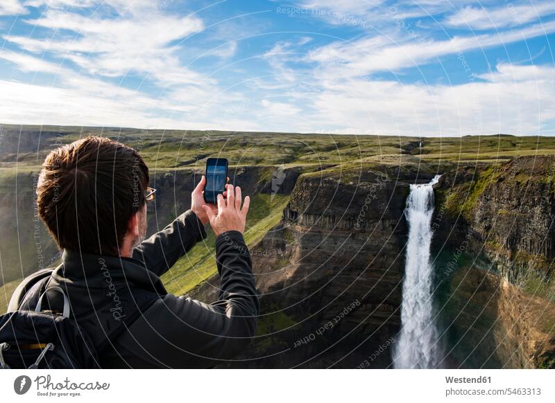 Wanderer, der den Haifoss-Wasserfall mit dem Handy fotografiert, während er in Island steht Farbaufnahme Farbe Farbfoto Farbphoto Außenaufnahme außen draußen