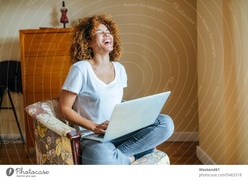 Glückliche junge Frau mit lockigem Haar sitzt im Sessel zu Hause mit Laptop sitzen sitzend weiblich Frauen Zuhause daheim Notebook Laptops Notebooks Locken