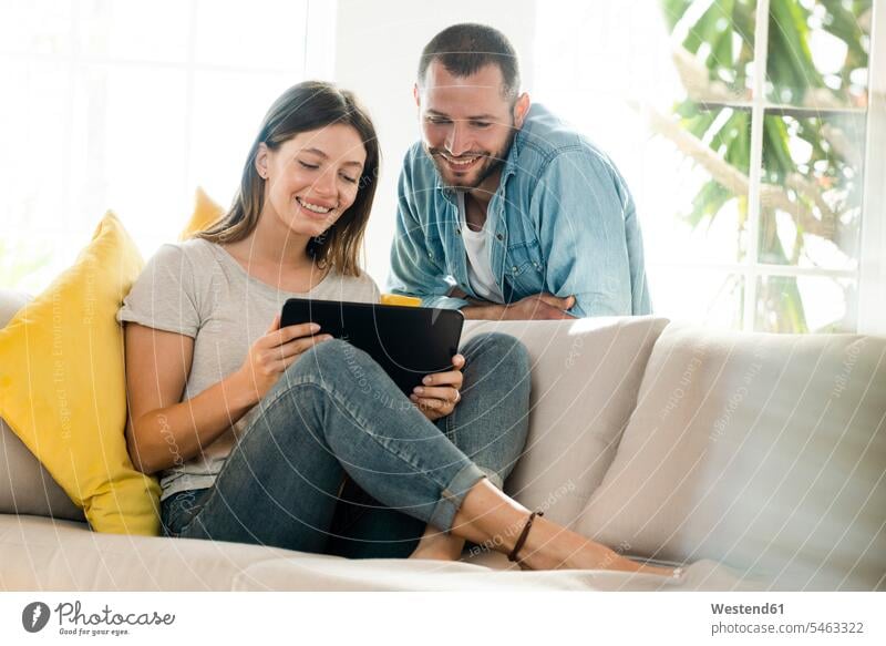 Glückliches Paar zu Hause im modernen Wohnzimmer auf der Couch und schaut gemeinsam auf ein Tablet Polster Couches Liege Sofas Medium sitzend sitzt entspannen
