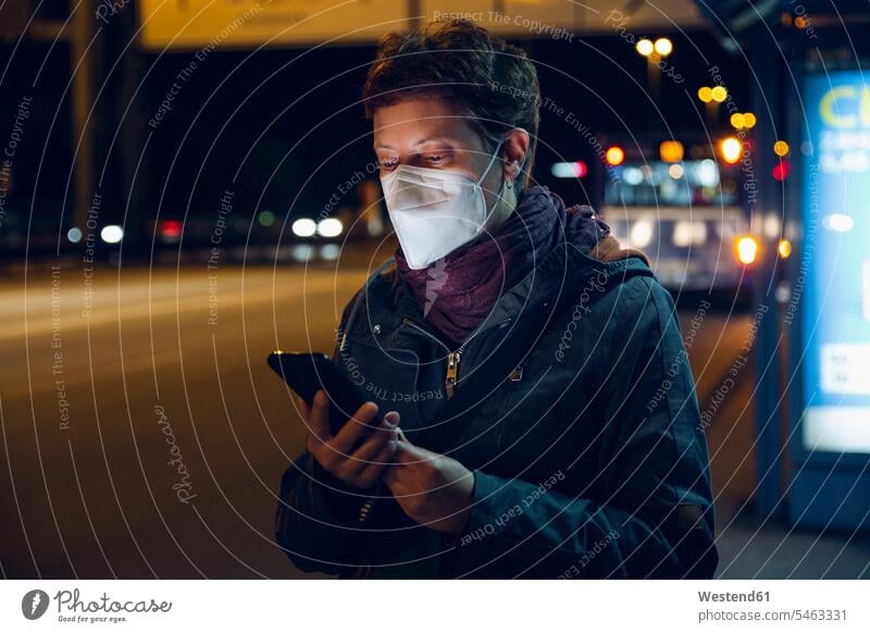 Frau mit Gesichtsschutzmaske, die ein Smartphone benutzt, während sie an der Bushaltestelle steht, während COVID-19 Farbaufnahme Farbe Farbfoto Farbphoto