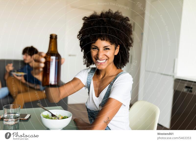 Porträt einer lächelnden Frau, die eine Bierflasche am Esstisch hebt sitzen sitzend sitzt Portrait Porträts Portraits Freundin Bierflaschen Tisch Tische