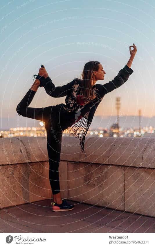 Spanien, Barcelona, Montjuic, junge Frau macht Yoga in der Abenddämmerung mit Stadt Lichter im Hintergrund stimmungsvoll Stimmung Dämmerung Abendstimmung