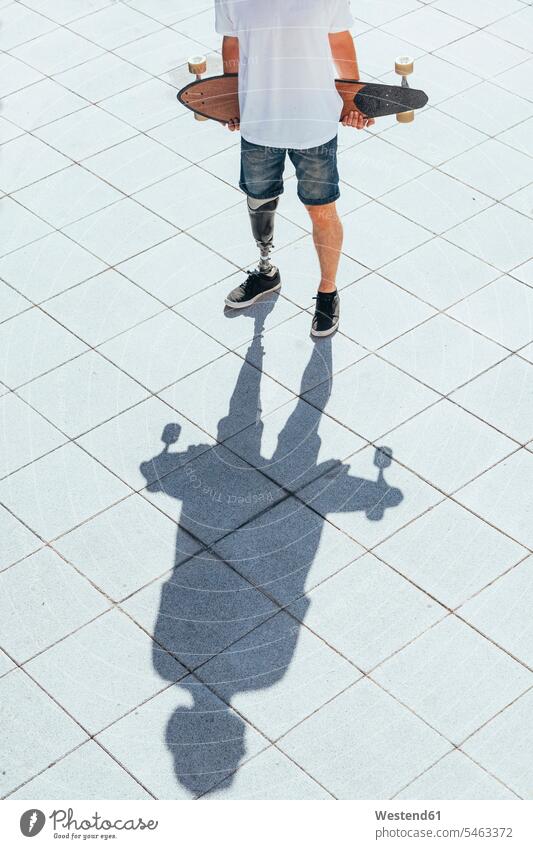Junger Mann mit Beinprothese hält Skateboard T-Shirts Jahreszeiten sommerlich Sommerzeit stehend steht Muße Individuell außen draußen im Freien staedtisch