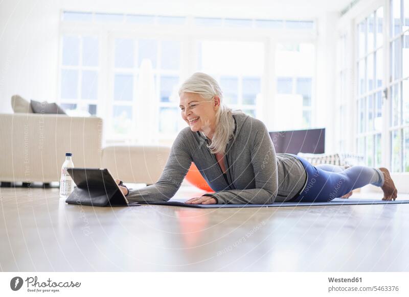 Lächelnde ältere Frau, die auf ein digitales Tablet schaut, während sie E-Learning-Übungen von einem digitalen Tablet in einer Wohnung macht Farbaufnahme Farbe