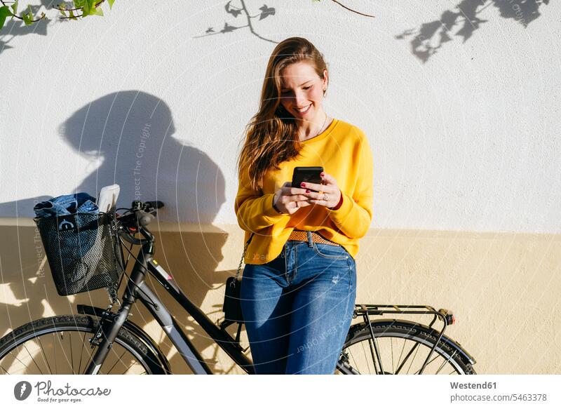 Lächelnde junge Frau mit Fahrrad mit Handy an einer Wand weiblich Frauen Wände Waende Bikes Fahrräder Räder Rad Mobiltelefon Handies Handys Mobiltelefone