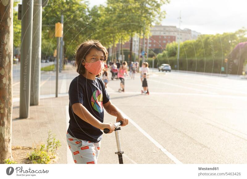 Porträt eines Jungen mit Schutzmaske beim Rollerfahren in der Stadt T-Shirts Kinderfahrzeuge Tretroller stehend steht Muße mobil geschützt schützen Absicherung