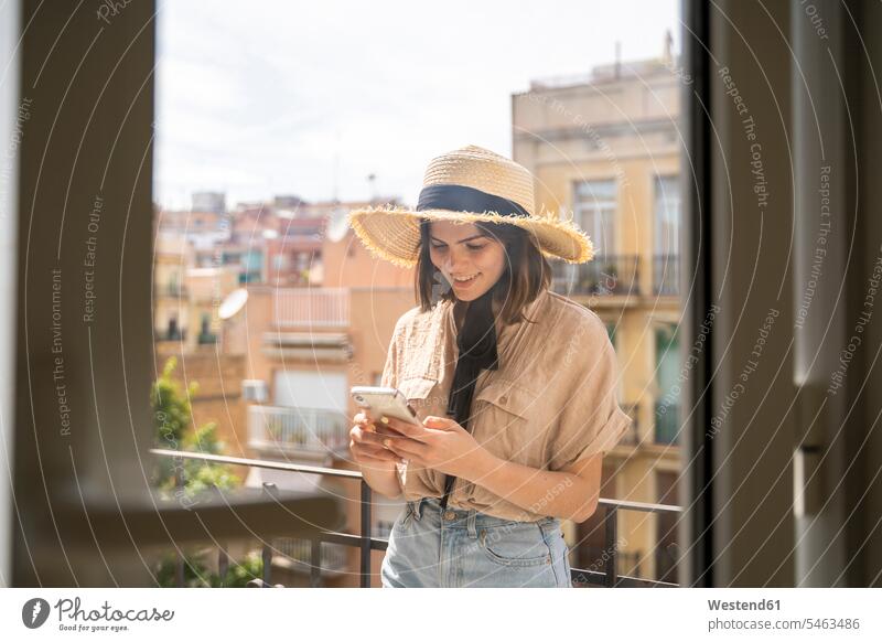 Lächelnde junge Frau mit Strohhut auf dem Balkon in der Stadt beim Telefonieren Hemden Telekommunikation telefonieren Handies Handys Mobiltelefon Mobiltelefone