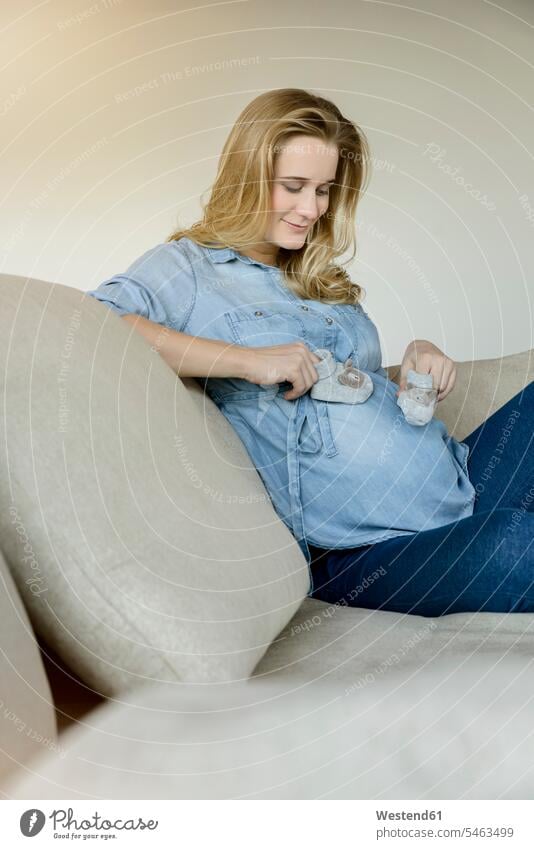Lächelnde schwangere Frau sitzt auf der Couch und hält Babyschuhe sitzen sitzend weiblich Frauen Schwangere halten Sofa Couches Liege Sofas Zuhause zu Hause