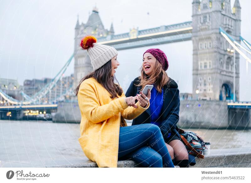 Zwei junge Touristen sitzen mit einem Smartphone an der Wand, mit der London Bridge im Hintergrund Bild Bilder Bildnis Foto Fotos Querformat Horizontalformat