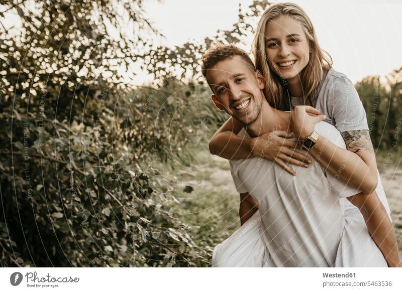 Porträt eines jungen lächelnden Paares, Mann trägt Frau auf dem Rücken T-Shirts Arm umlegen Umarmung Umarmungen abends freuen Frohsinn Fröhlichkeit Heiterkeit