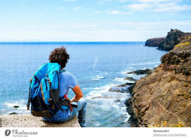 Spanien, Kanarische Inseln, Gran Canaria, Mann mit Rucksack auf Klippe sitzend Tourist Touristen Aussicht Ausblick Ansicht Überblick Reisende Reisender Männer