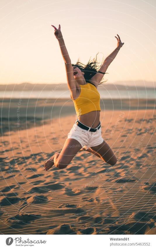 Glückliche Teenagerin springt bei Sonnenuntergang am Strand in die Luft Beach Straende Strände Beaches Sonnenuntergänge junges Mädchen Teenagerinnen weiblich